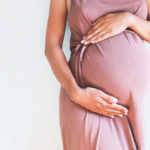 Pregnant woman in dress holds hands on belly on a white background.